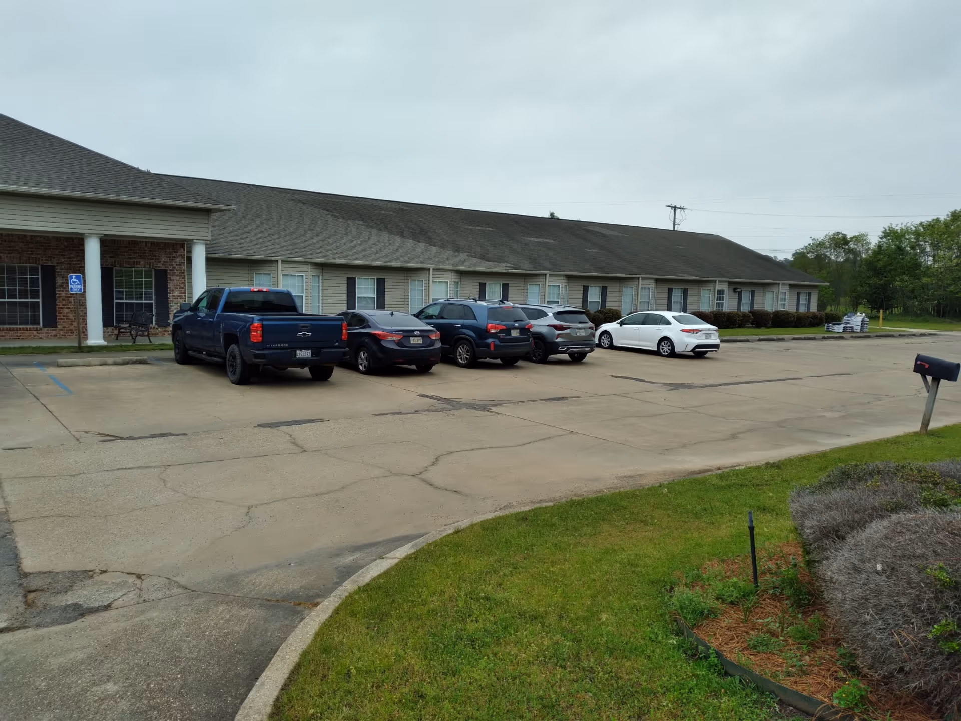 Parking lot in front of a single-story building with several cars parked. The building has a brick and siding exterior with white columns near the entrance. There is a grassy area with some shrubs and a mailbox in the foreground.