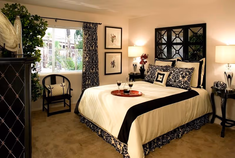 A well-decorated bedroom with a large bed featuring black and white patterned pillows and bedding. There is a red tray with two glasses of wine and a wine bottle on the bed. The room has beige carpet, two bedside tables with lamps, framed artwork on the wall, a window with patterned curtains, a black chair with a cushion, and a black dresser with decorative items.