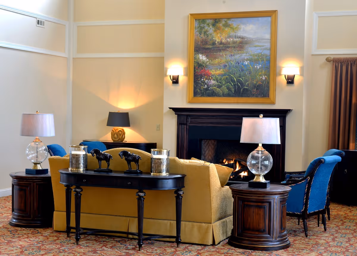 A cozy living room area with a yellow sofa facing a lit fireplace. Two blue armchairs are positioned on either side of the sofa. There are two round wooden side tables with glass lamps on each side of the sofa. A black console table behind the sofa holds decorative horse figurines and candles. Above the fireplace hangs a large framed painting of a nature scene with flowers and water. The room has warm lighting and a patterned carpet.