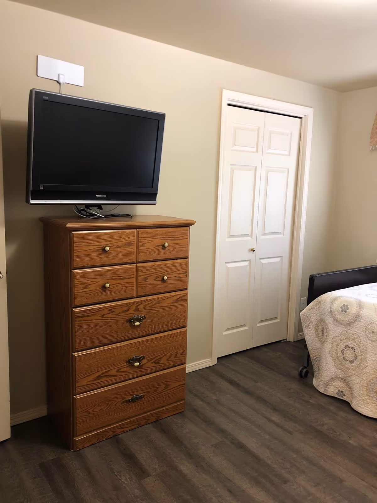 A bedroom corner with a wooden dresser featuring six drawers and a flat-screen TV mounted on the wall above it. To the right, there is a white closet door and part of a bed with a patterned quilt visible. The floor is dark wood, and the walls are light-colored.