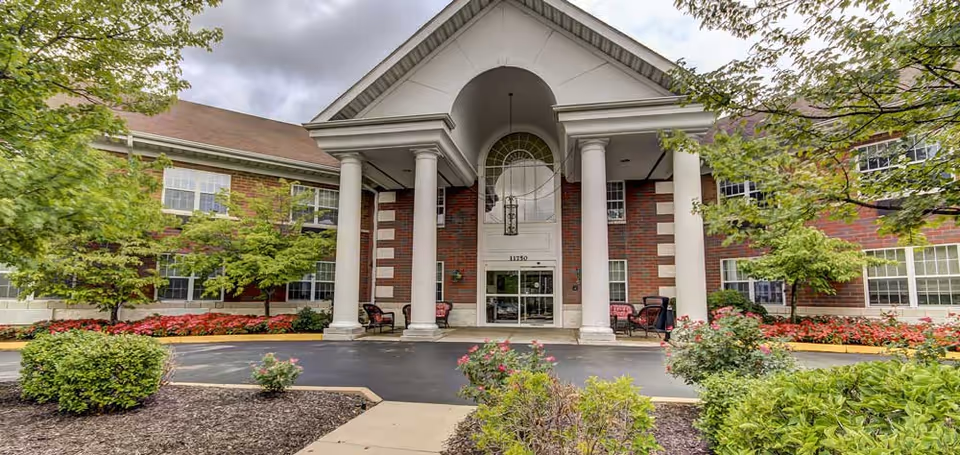 Front exterior view of a senior living facility named The Estates of Hidden Lake, featuring a brick building with white columns at the entrance, surrounded by landscaped bushes, flowers, and trees under a partly cloudy sky.