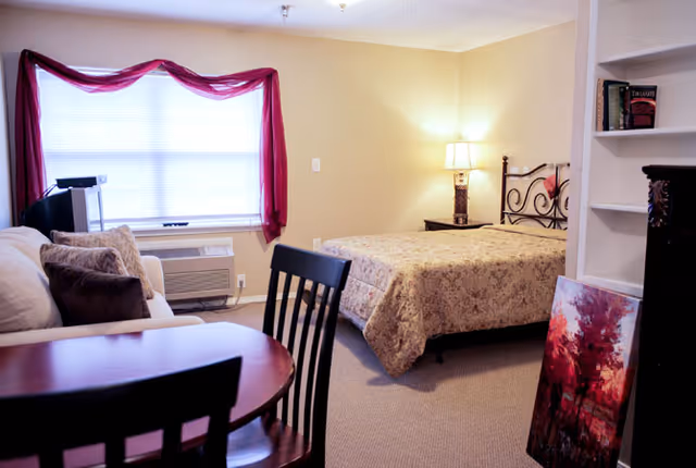 A cozy bedroom with a bed covered in a patterned quilt, a bedside table with a lamp, a window with red curtains, a small sofa with cushions, a round wooden table with chairs, and a painting leaning against a white bookshelf.