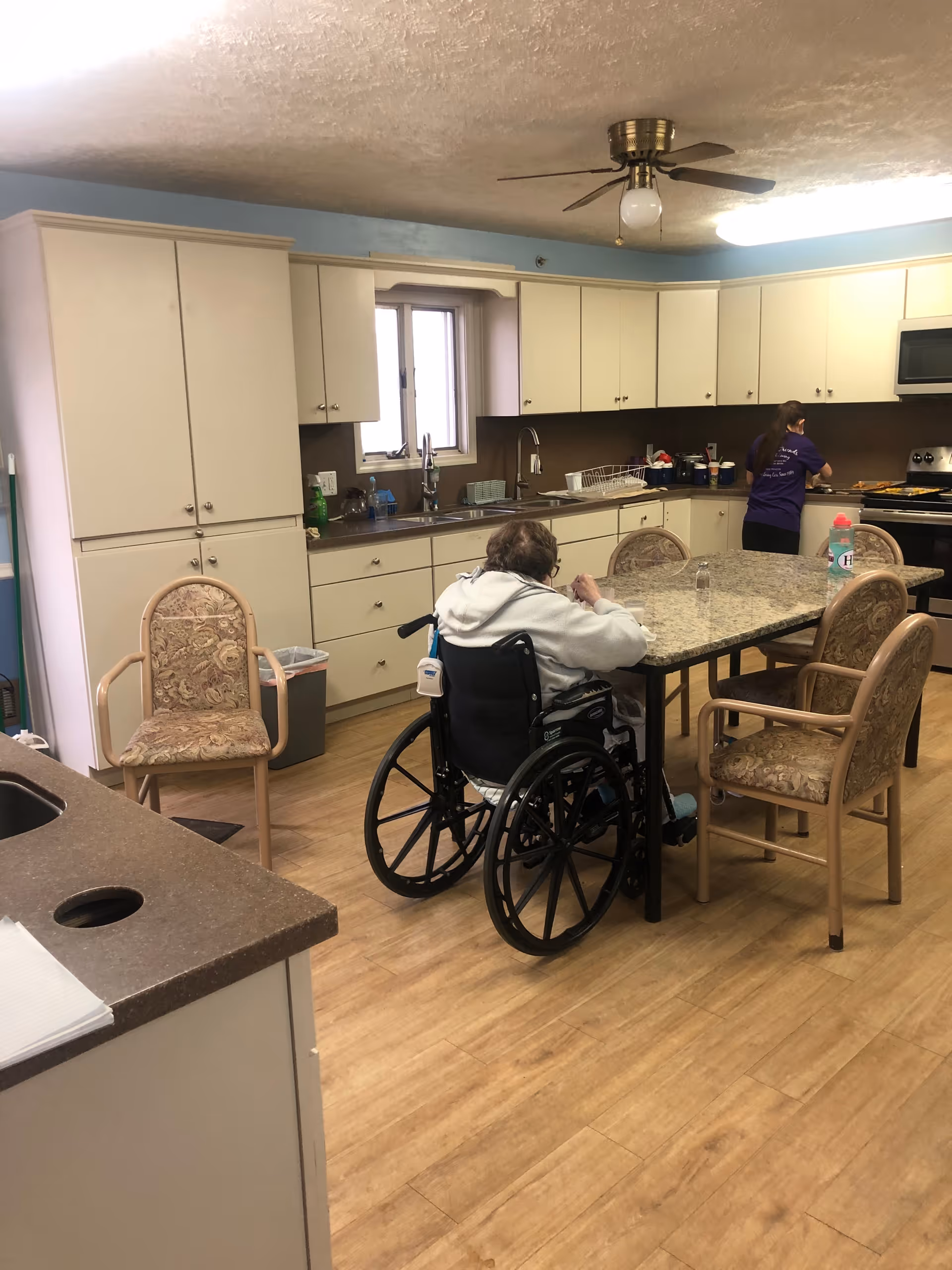 A kitchen area in an assisted living facility with white cabinets, a ceiling fan, and a granite table surrounded by chairs. An elderly person in a wheelchair is seated at the table, and a staff member is preparing food at the stove in the background.