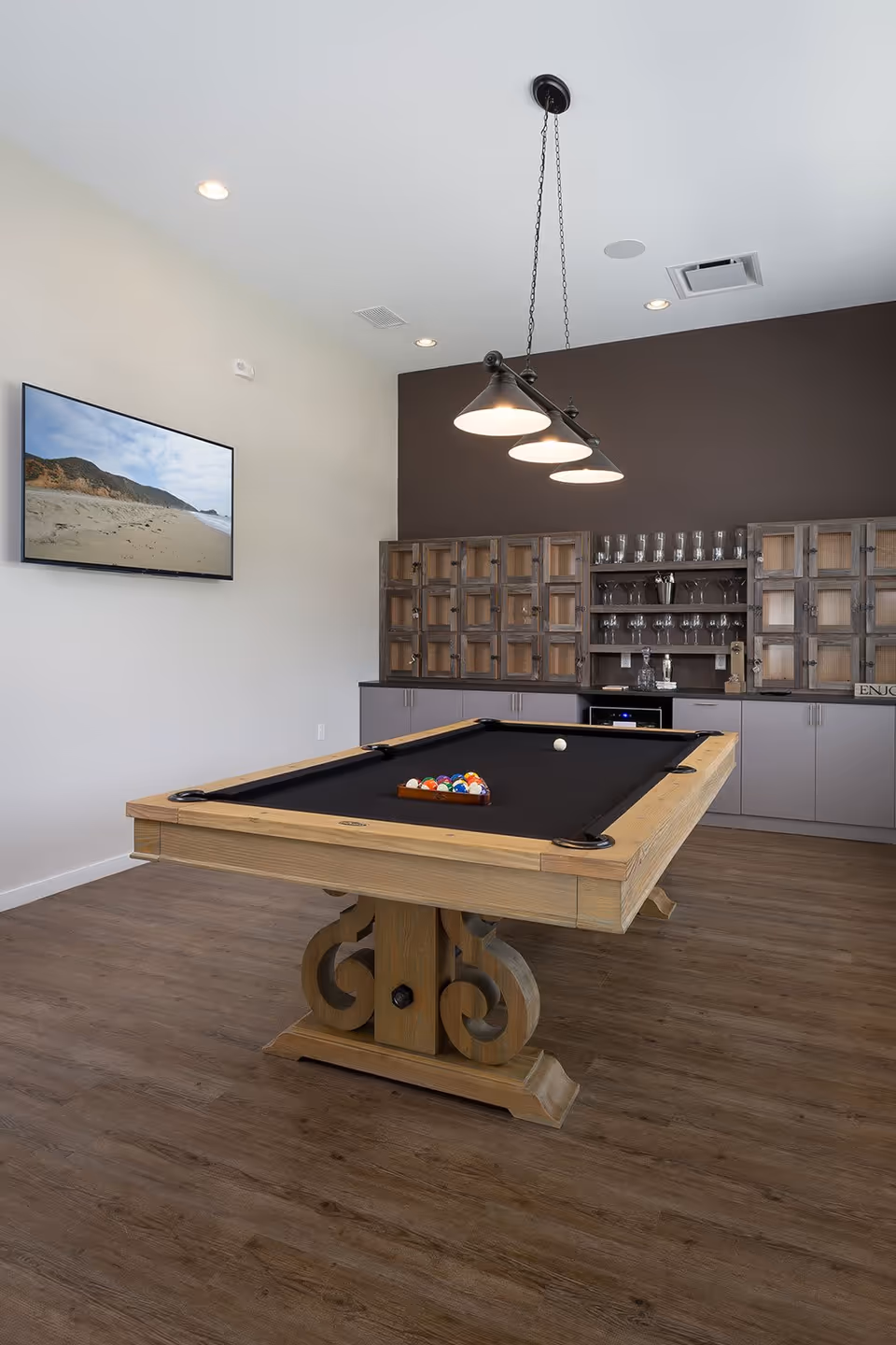 A modern game room featuring a wooden pool table with a black felt surface, a set of pool balls arranged in a triangle, and a single white cue ball. Above the table hangs a three-light pendant fixture. In the background, there is a wall with a dark brown accent and cabinetry with glass doors displaying glassware. A flat-screen TV mounted on a white wall shows a beach scene.