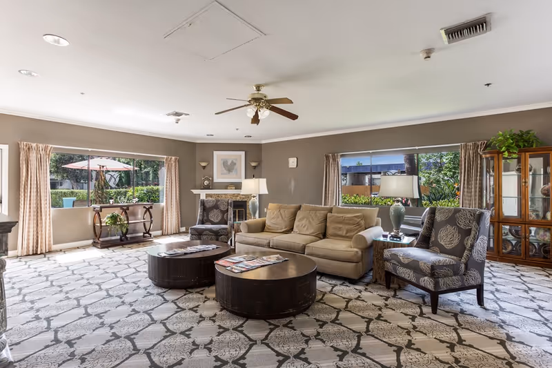Bright communal living room with a sofa, patterned armchairs, round coffee tables, large windows, and a display cabinet.