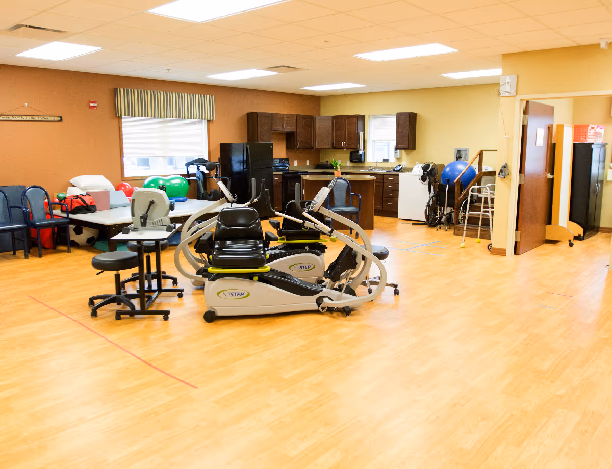 Spacious interior activity/therapy room with exercise equipment, chairs, and a small kitchenette in a senior living facility.