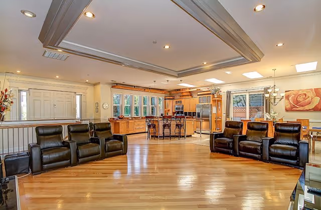 Spacious open-concept living area with polished hardwood floors, two sets of black leather recliners arranged facing each other, and a large kitchen in the background featuring wooden cabinetry, stainless steel appliances, and a kitchen island with bar stools. The room is well-lit with recessed ceiling lights and natural light from windows and a glass door leading outside.