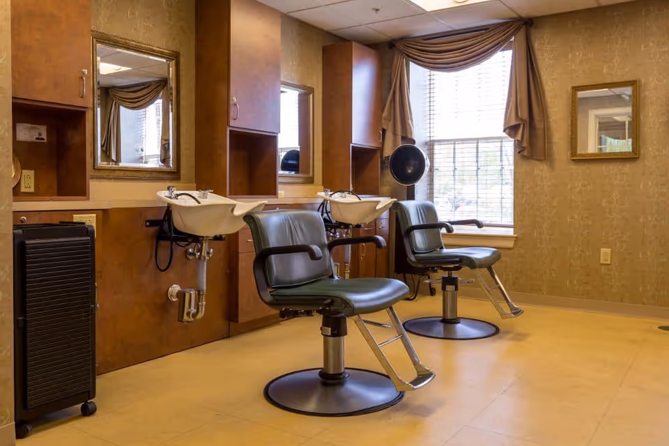 Interior view of a hair salon area with two green salon chairs in front of two white wash basins. There are mirrors mounted on the wall above the basins, wooden cabinets, a window with brown curtains, and a hair dryer attached to the wall.