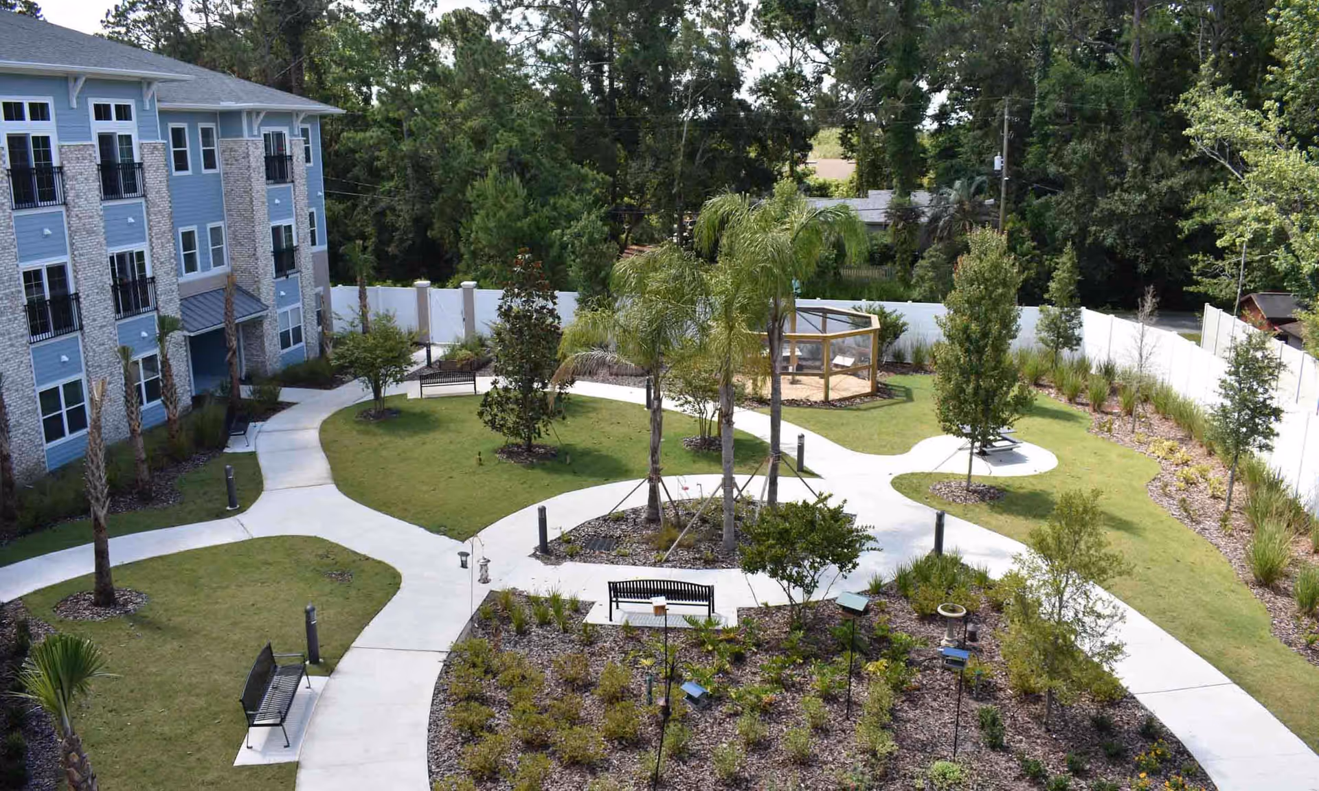 Outdoor garden area at HarborChase of Mandarin featuring winding concrete pathways, green lawns, various trees including palm trees, benches for seating, and a fenced enclosure in the background surrounded by a white privacy fence and dense trees.