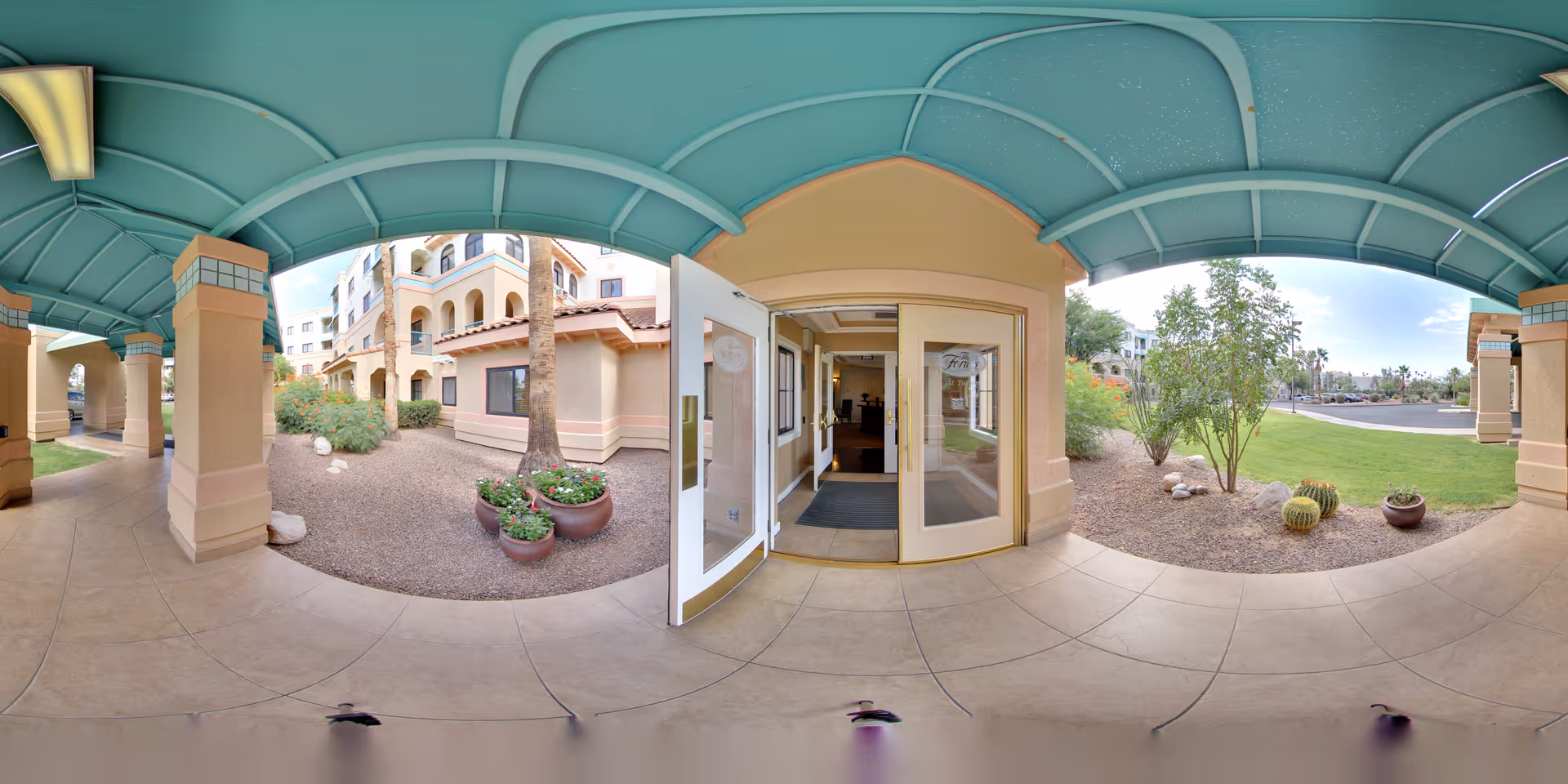 Covered outdoor walkway with a teal arched ceiling leading to the entrance of a building. The entrance has double glass doors, one of which is open, showing a glimpse of the interior. Surrounding the walkway are desert landscaping elements including rocks, potted plants, cacti, and small trees. The building exterior is beige with architectural details and multiple windows.