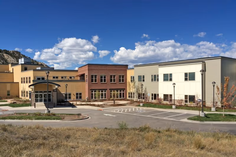 Exterior view of Castle Peak Assisted Living facility showing a modern building with multiple sections in beige, red brick, and light gray colors under a blue sky with scattered clouds. There is a parking area with marked spaces and street lamps in front of the building.