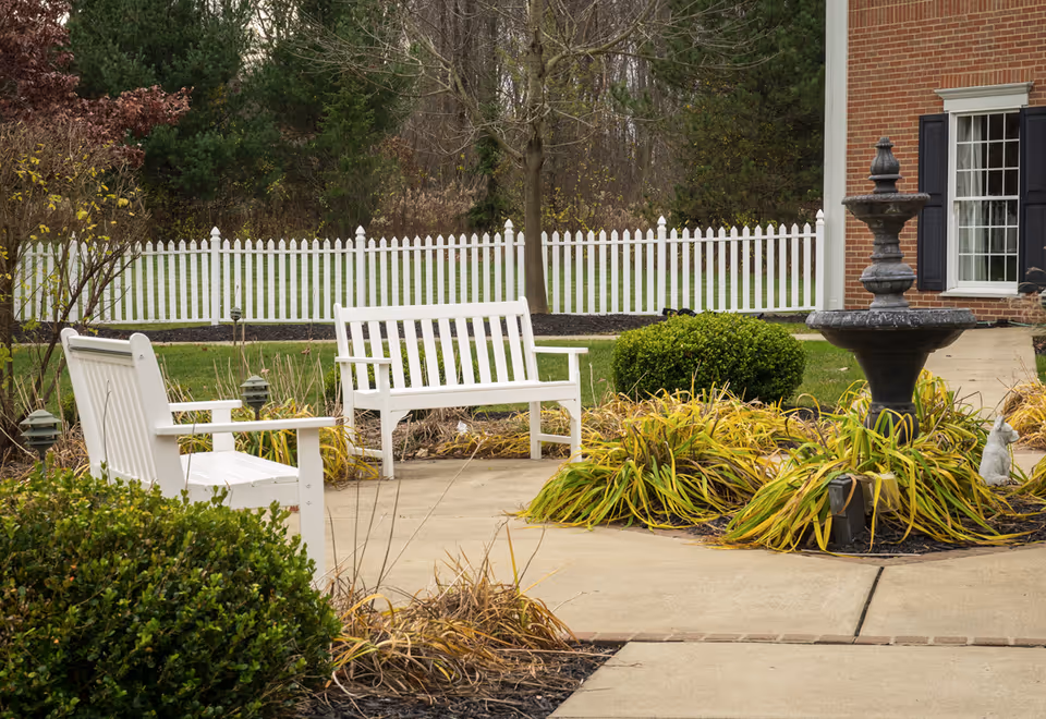 Outdoor courtyard with white benches, a fountain, walkways, shrubs, and a white picket fence by a brick building.