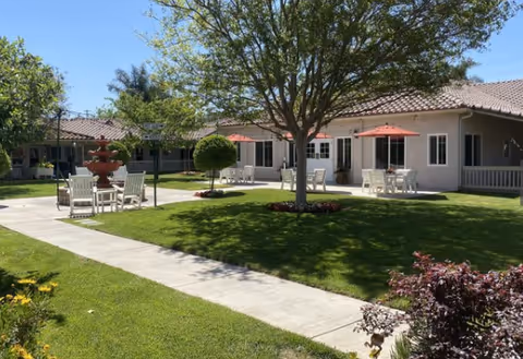 Outdoor courtyard area at The Gardens of Riverside with green grass, a large tree in the center, several white tables and chairs with red umbrellas, a red fountain, and a building with a tiled roof in the background under a clear blue sky.