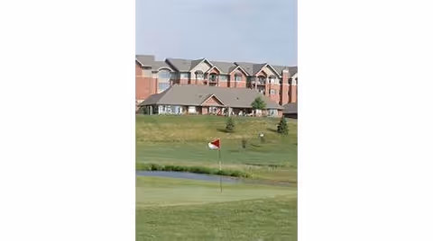 View of a golf course green with a red and white flag in the foreground, a small pond behind it, and a large multi-story brick building with a gray roof in the background under a cloudy sky.