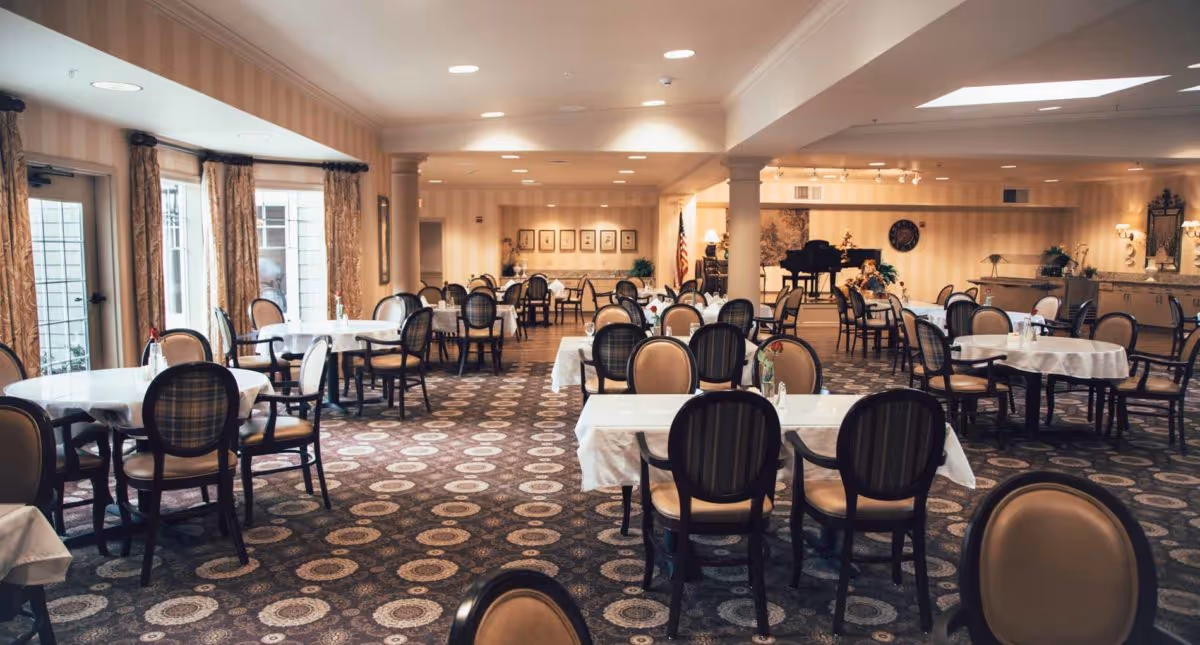 A spacious dining room with multiple round and rectangular tables covered with white tablecloths, surrounded by cushioned chairs. The room has patterned carpet flooring, large windows with curtains, and soft overhead lighting. In the background, there is a grand piano and decorative wall art.