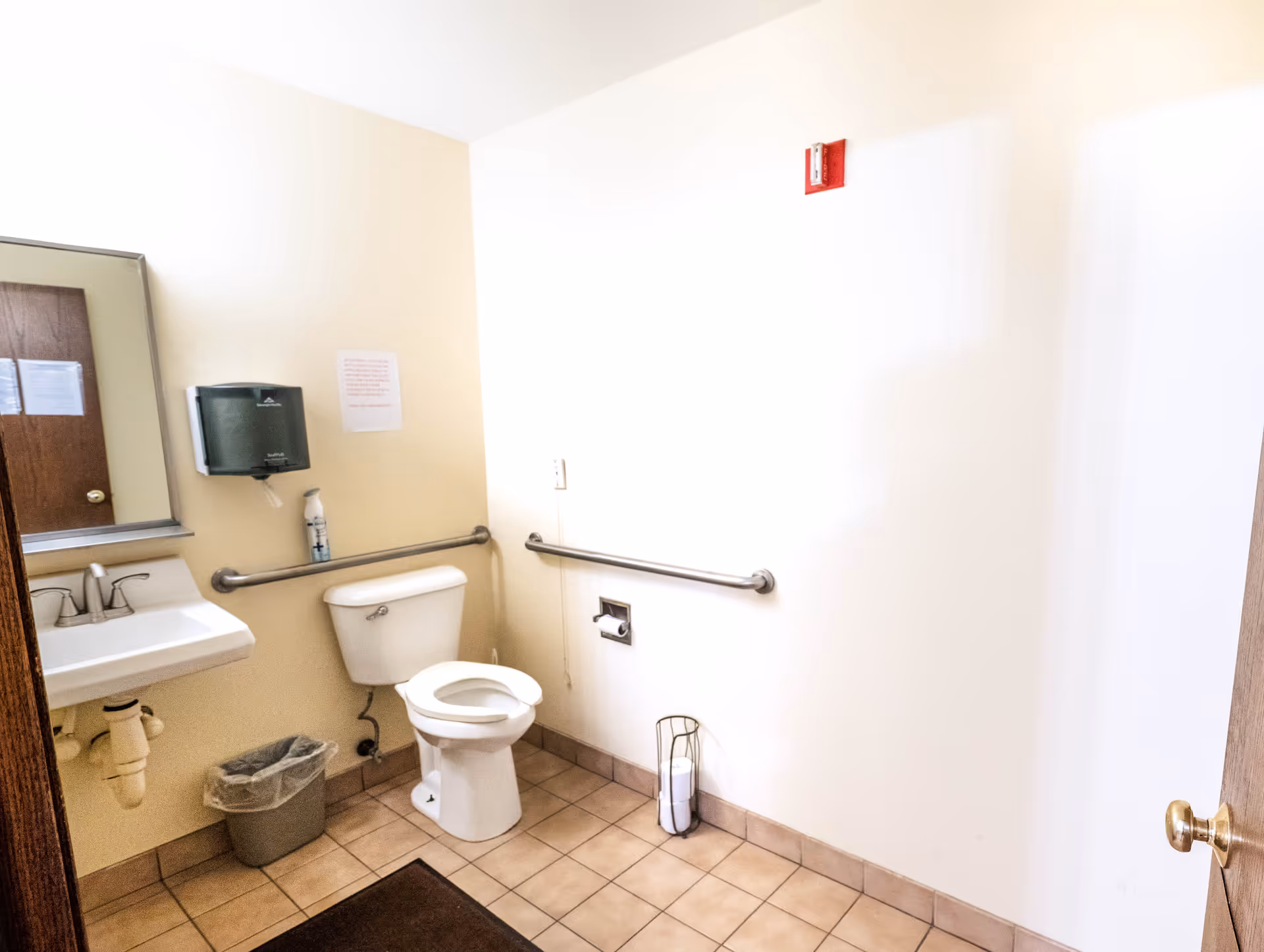 A clean and accessible bathroom with a white toilet, a sink with a mirror above it, a trash can, and grab bars on the walls. The floor is tiled and there is a paper towel dispenser mounted on the wall.