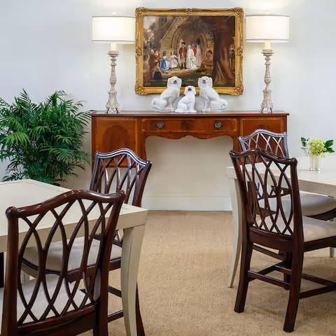 A dining area with wooden chairs featuring intricate back designs around beige tables. Against the wall is a wooden sideboard with three white ceramic dog figurines and two table lamps with white shades. Above the sideboard hangs a framed painting depicting a historical scene. A green potted plant is visible to the left of the sideboard.