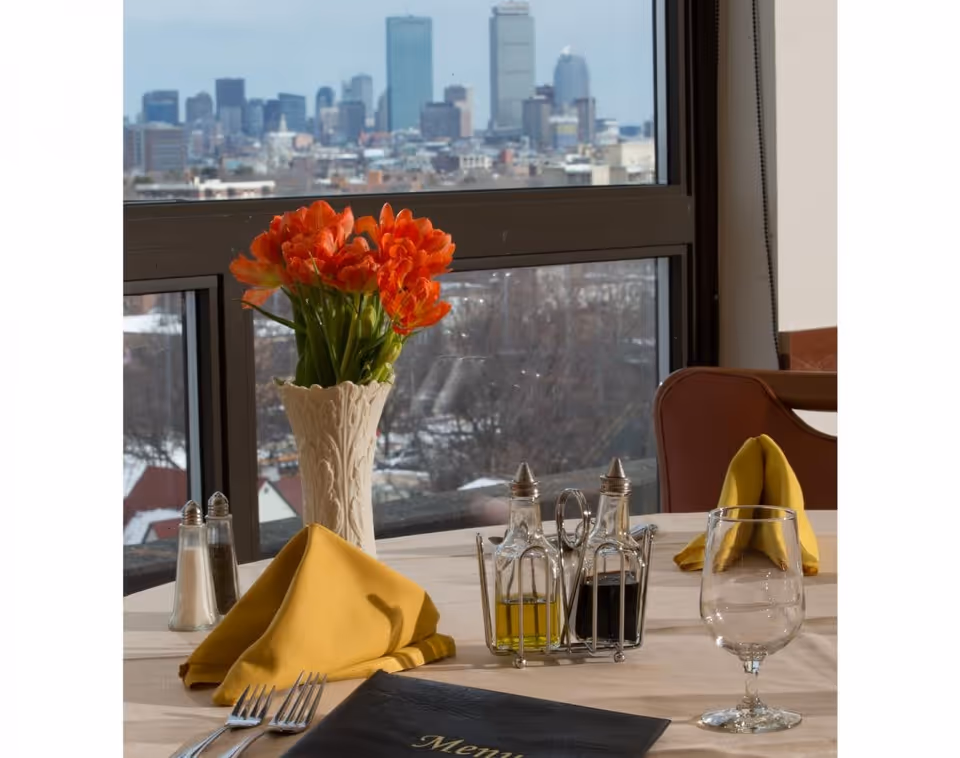 A dining table set with yellow folded napkins, silverware, a glass, salt and pepper shakers, and a condiment set with oil and vinegar bottles. A white vase with bright orange flowers is placed near a large window showing a city skyline in the background.