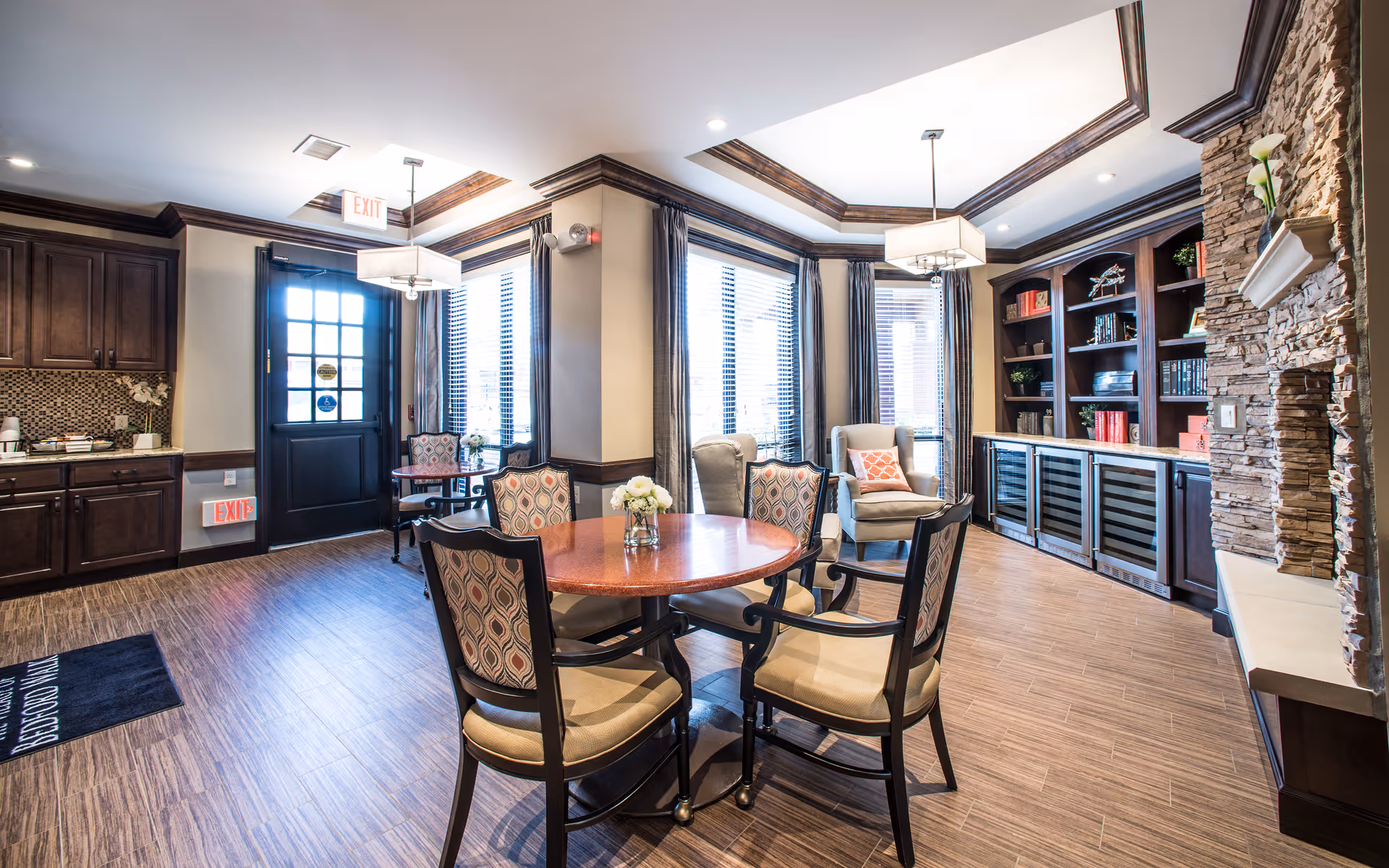 A bright and inviting common area with a round wooden table surrounded by four cushioned chairs. The room features large windows with blinds and curtains, two armchairs with a decorative pillow, built-in dark wood bookshelves filled with books and decor, and a stone fireplace with a mantel. The floor is wood-patterned, and there is a black door with exit signs above and below it. The ceiling has recessed lighting and two hanging light fixtures.