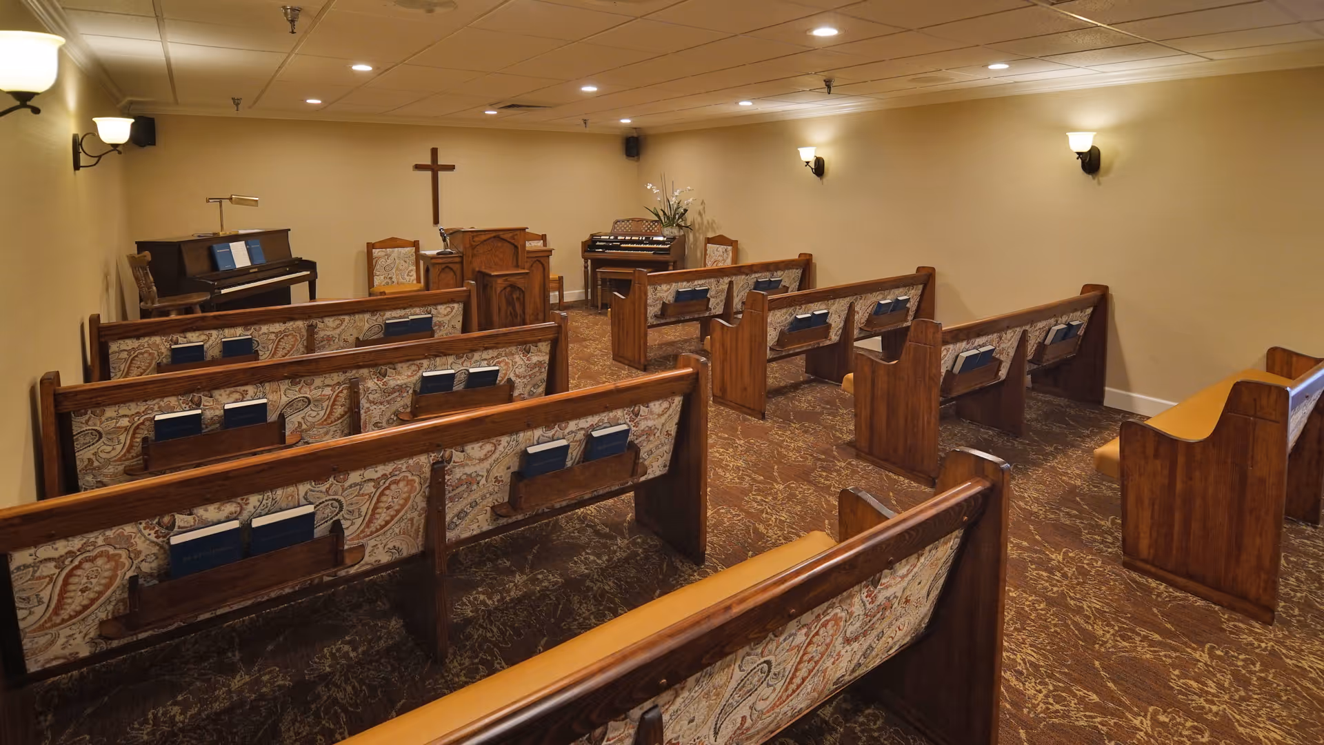 Interior view of a small chapel or worship room with wooden pews arranged in rows facing a wooden pulpit. There is a cross mounted on the wall behind the pulpit, a piano on the left side, and an organ on the right side. The room has beige walls, carpeted floor, and wall-mounted lights.