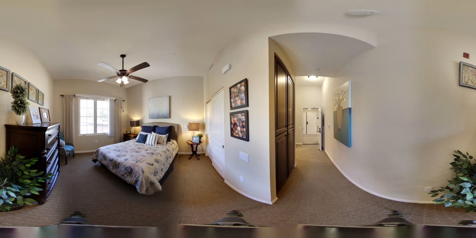 A well-lit bedroom with a large window, a ceiling fan with lights, a bed with floral bedding and multiple pillows, a wooden dresser with framed pictures and a plant on top, a small side table with a lamp and a photo frame, and a hallway leading to a door. The walls are decorated with framed artwork and the floor is carpeted.