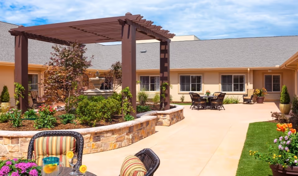 Outdoor courtyard area of a senior living facility with a stone planter bed, a wooden pergola, a water fountain, and several seating areas with tables and chairs. The courtyard is surrounded by a beige building with multiple windows and a clear blue sky overhead.