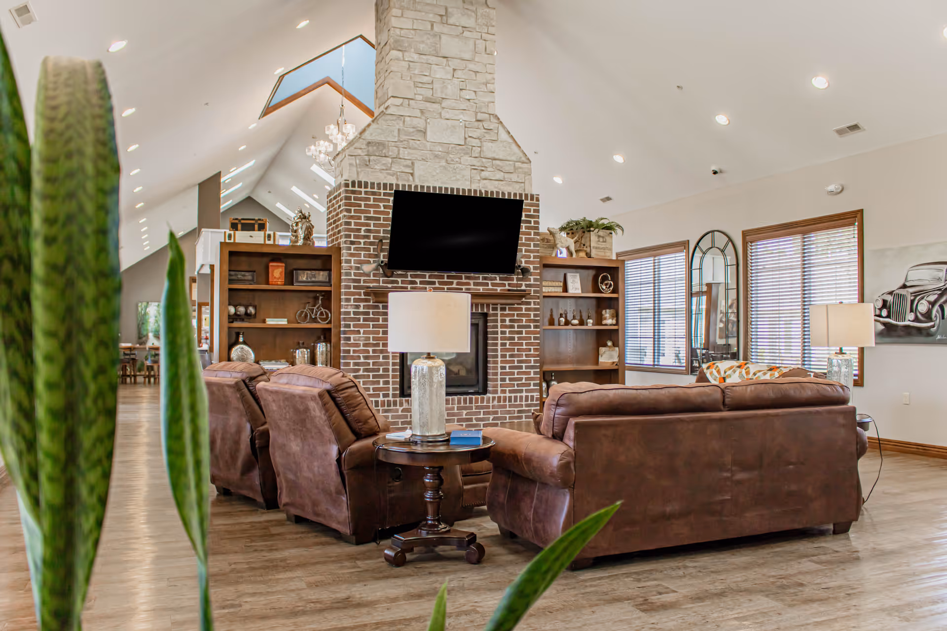 A spacious living room area with brown leather sofas and armchairs arranged around a brick and stone fireplace with a mounted flat-screen TV above it. The room features wooden bookshelves on either side of the fireplace, large windows with blinds, and decorative lamps on side tables. The ceiling is vaulted with recessed lighting and a chandelier visible in the background. A tall green plant is partially visible in the foreground.