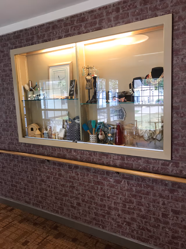 A glass display case mounted on a brick wall with various decorative items inside, including small mannequins with jewelry, kitchen utensils, glassware, and a small wooden birdhouse. The display is illuminated from above and reflects windows and outdoor scenery.