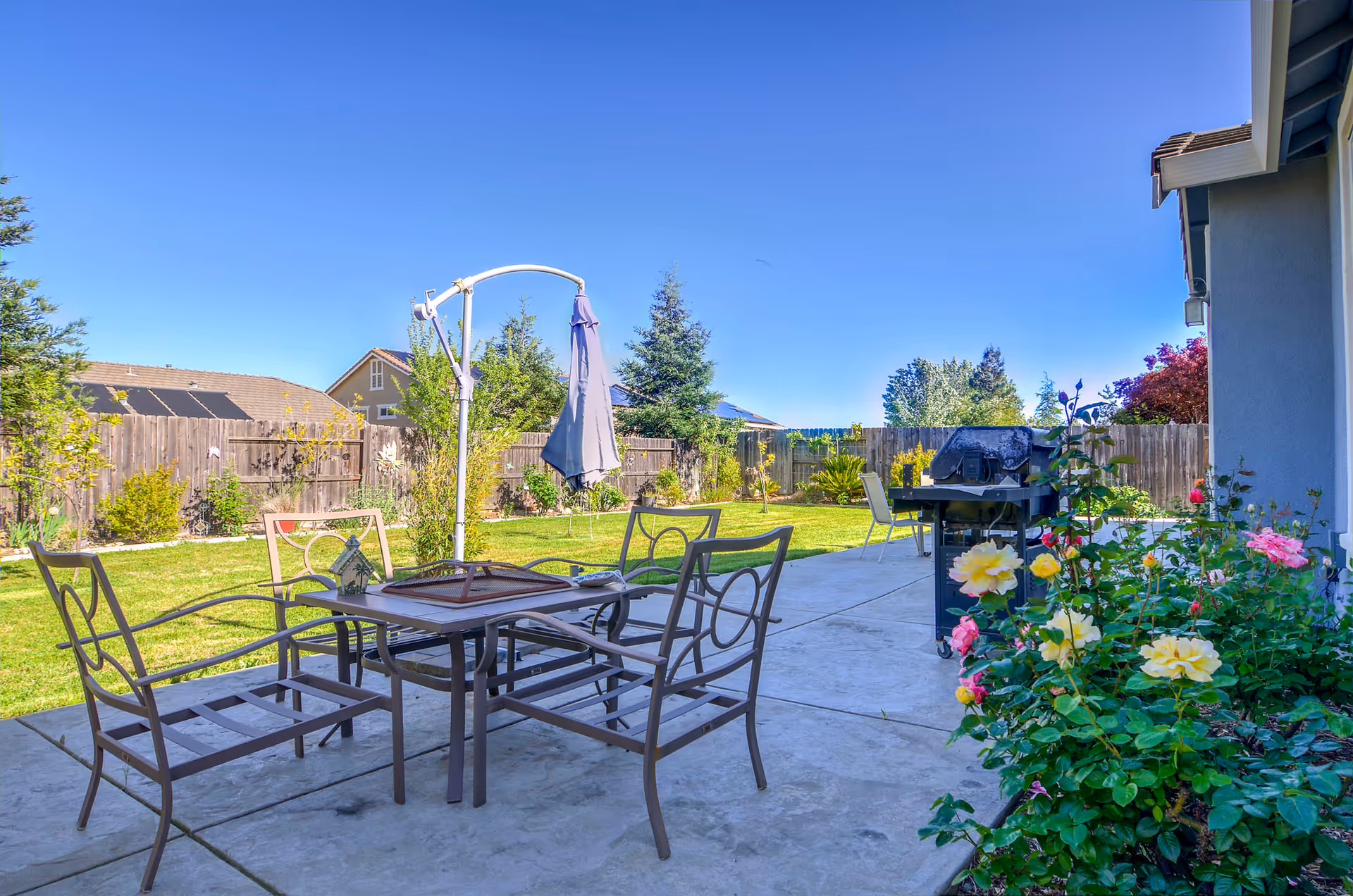 Outdoor patio area with a metal table and four chairs on a concrete surface, a closed umbrella, a barbecue grill, and a garden with green grass, flowers, and trees under a clear blue sky.