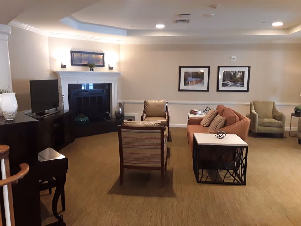 A cozy living room area with a brown sofa, two armchairs, a striped chair, and a small table with decorative items. There is a fireplace with a mantle, a flat-screen TV on a stand, and framed pictures on the wall. The room has beige walls and carpeted floor with soft lighting.