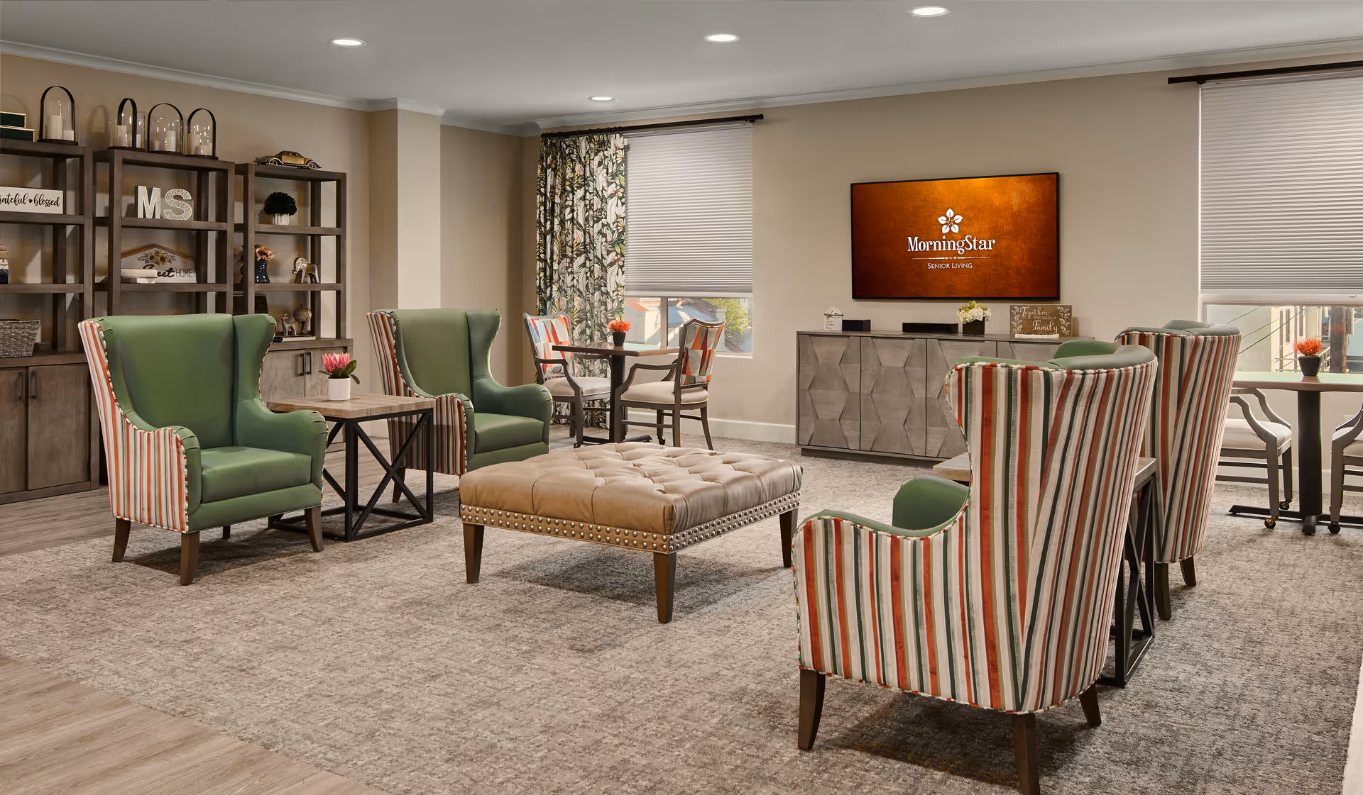 A senior living facility common area with several upholstered armchairs, some with green cushions and others with striped fabric. There is a large tufted ottoman in the center on a carpeted floor. In the background, there are windows with blinds and floral curtains, a wooden shelving unit with decorative items, and a wall-mounted TV displaying the MorningStar Senior Living logo. Small tables with potted plants are placed near the chairs.