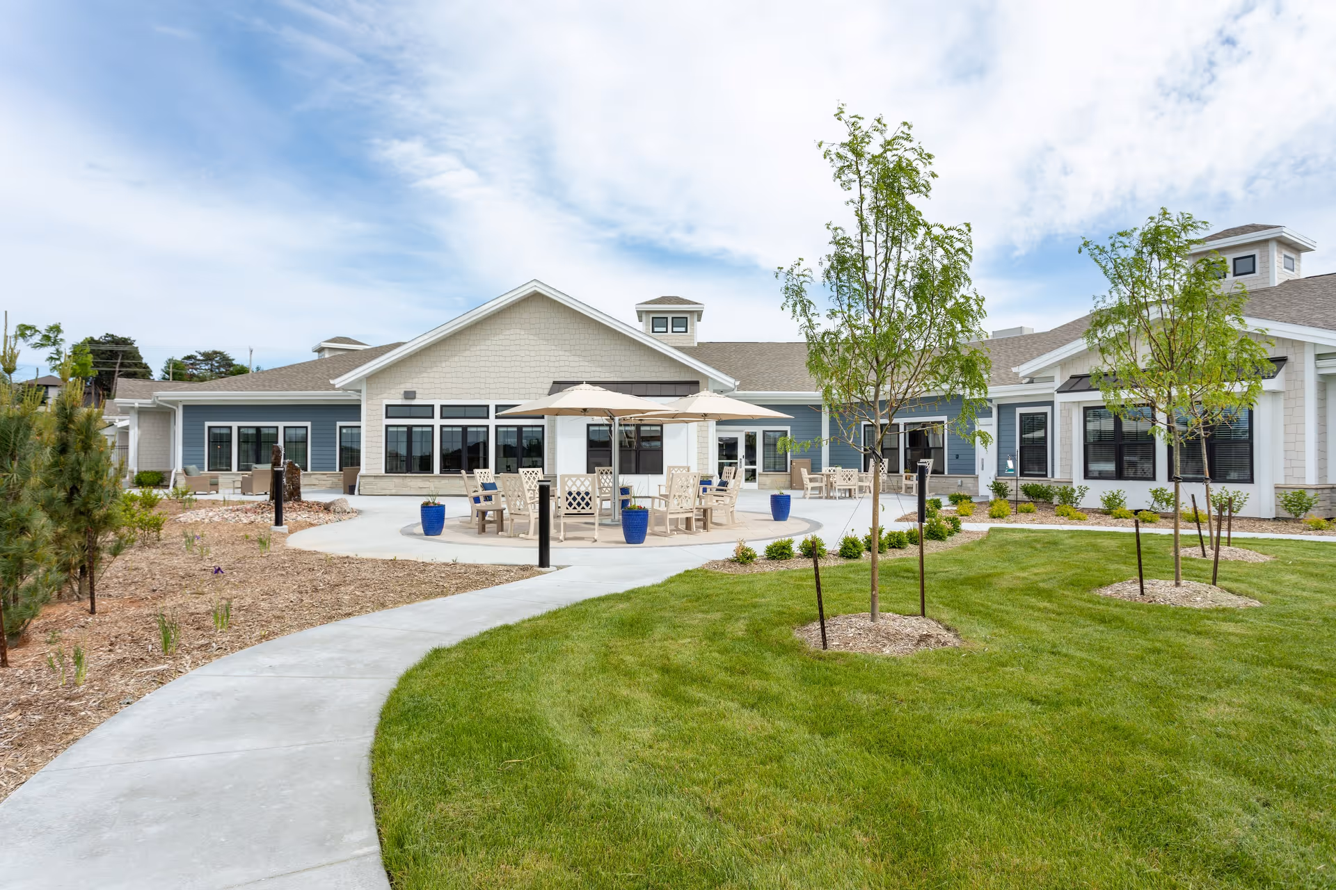 Outdoor patio area of CountryHouse Residence featuring a curved concrete walkway, green lawn with young trees, and seating areas with tables and umbrellas in front of a single-story building with large windows under a partly cloudy sky.