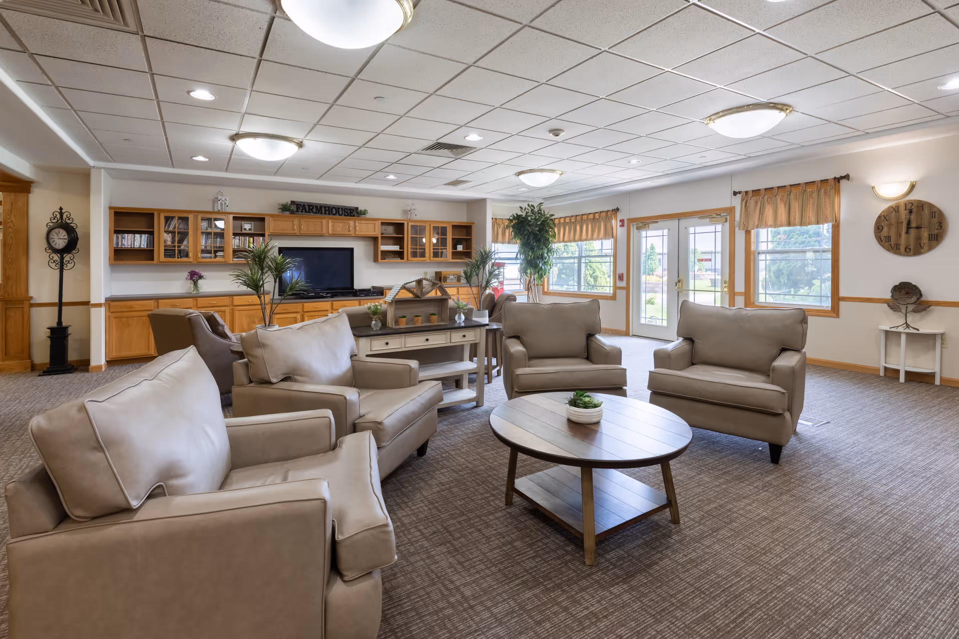 Spacious communal living room with beige armchairs around a round coffee table, built-in cabinets and large windows.