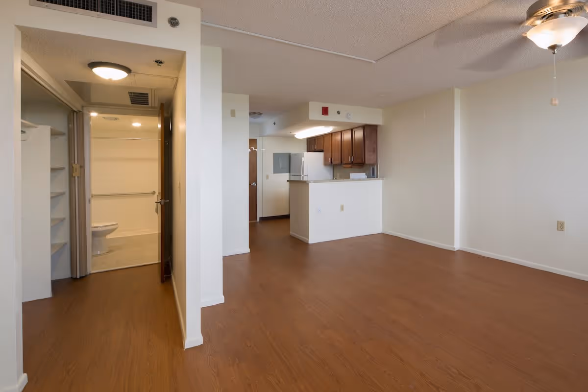 Empty open living room with wood floors, a kitchenette with upper cabinets, and a bathroom entrance on the left.