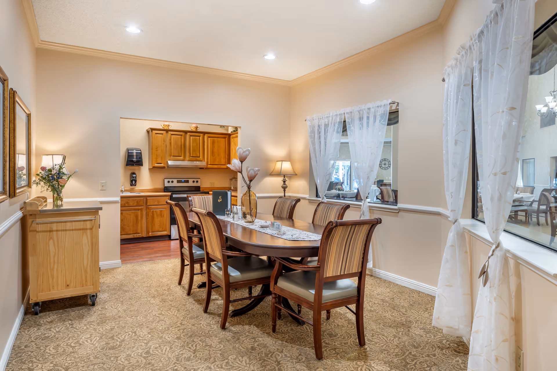 A dining room with a wooden table and six chairs, a floral centerpiece, and an open pass-through to a small kitchen.