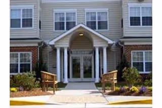 Entrance of a residential building with a covered porch supported by white columns, two wooden benches on either side of the walkway, and landscaped bushes and flowers along the front.