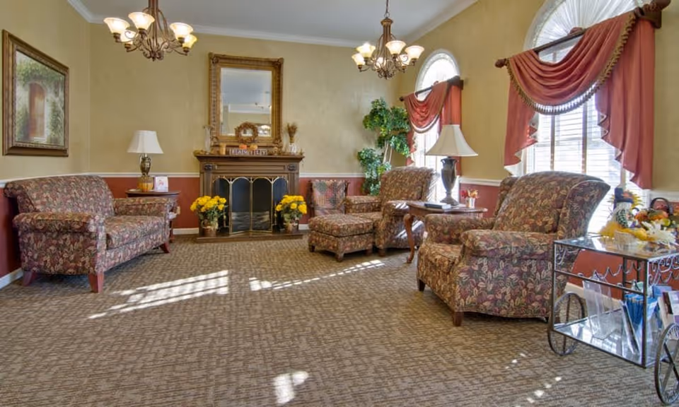 A cozy senior living facility common area with floral upholstered armchairs and a loveseat arranged around a fireplace with a large mirror above it. The room has beige walls with a red lower border, two chandeliers, table lamps, framed artwork, and windows with red drapes letting in natural light. A small metal cart with brochures and a fruit basket is also visible.