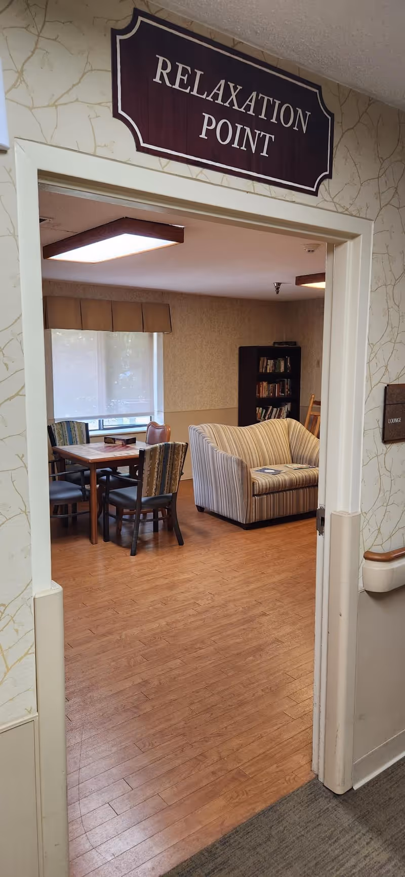 View through a doorway into a room labeled 'Relaxation Point' with a wooden floor, a striped couch, a bookshelf, and a table with chairs near a window with blinds.