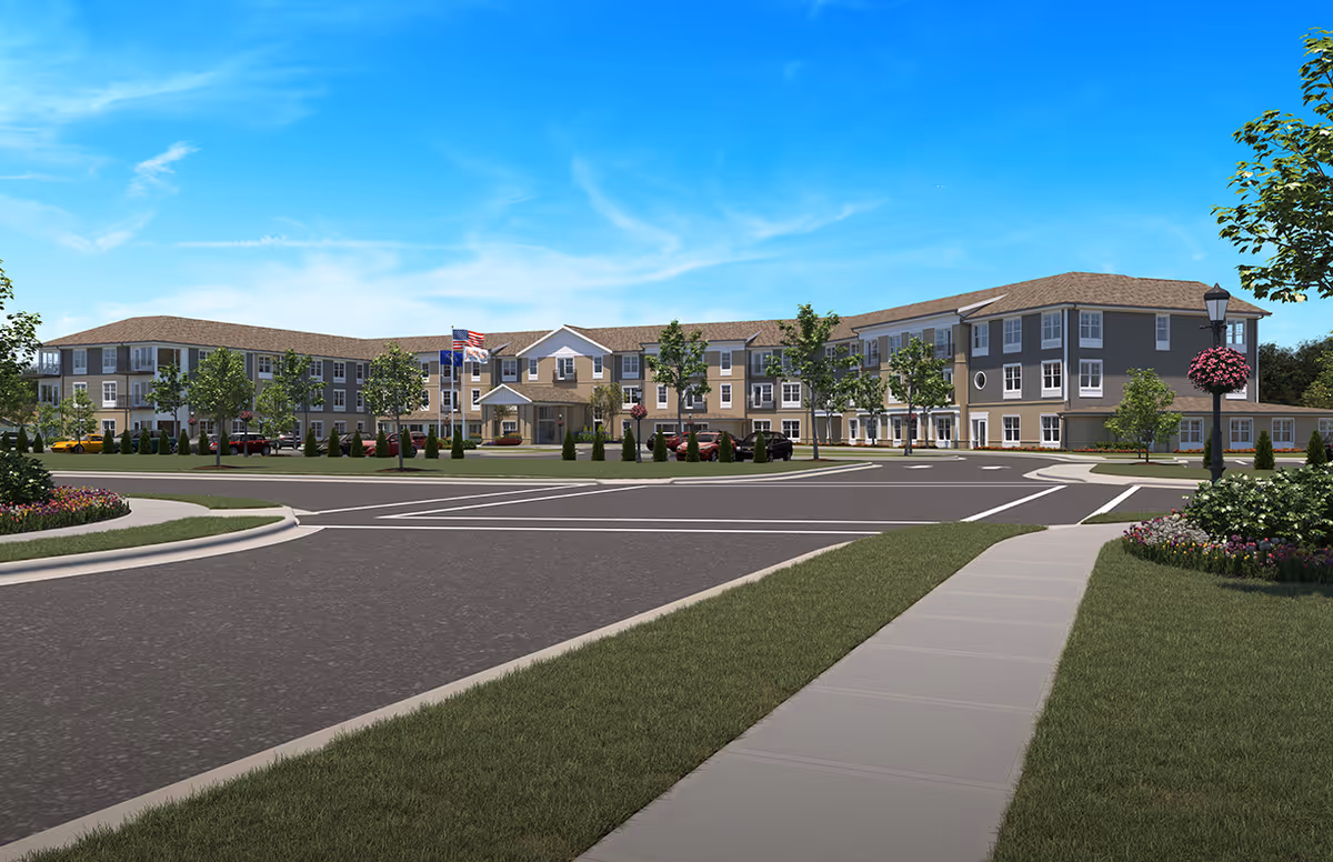 Exterior view of a large, modern three-story senior living facility building with beige and gray siding, multiple windows, and a central entrance with a covered porch. The building is surrounded by landscaped greenery, trees, and a paved road with sidewalks. An American flag and another flag are visible near the entrance under a clear blue sky.
