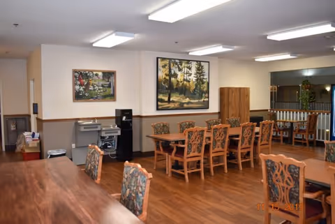 Interior view of a dining room in Orchards Assisted Living featuring wooden tables and chairs with floral upholstery. The room has hardwood floors, two large framed paintings on the wall, a water cooler, and a small counter area with a sink. The ceiling has fluorescent lighting and there is a railing visible in the background.