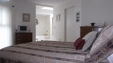 A bedroom with a neatly made bed featuring patterned bedding and pillows. There is a wooden dresser with a small TV and decorative items on top. The room has white walls and a doorway leading to a bathroom with a visible toilet and a wreath on the wall.