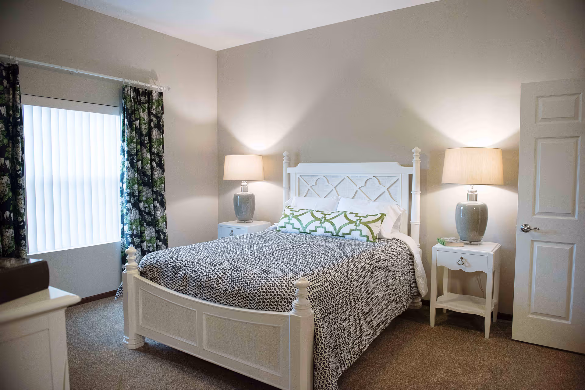 A cozy bedroom with a white wooden bed frame, a patterned gray and white bedspread, and two green and white decorative pillows. There are two matching white nightstands on either side of the bed, each with a gray lamp. A window with floral curtains is on the left wall, and a white door is partially open on the right side of the room.