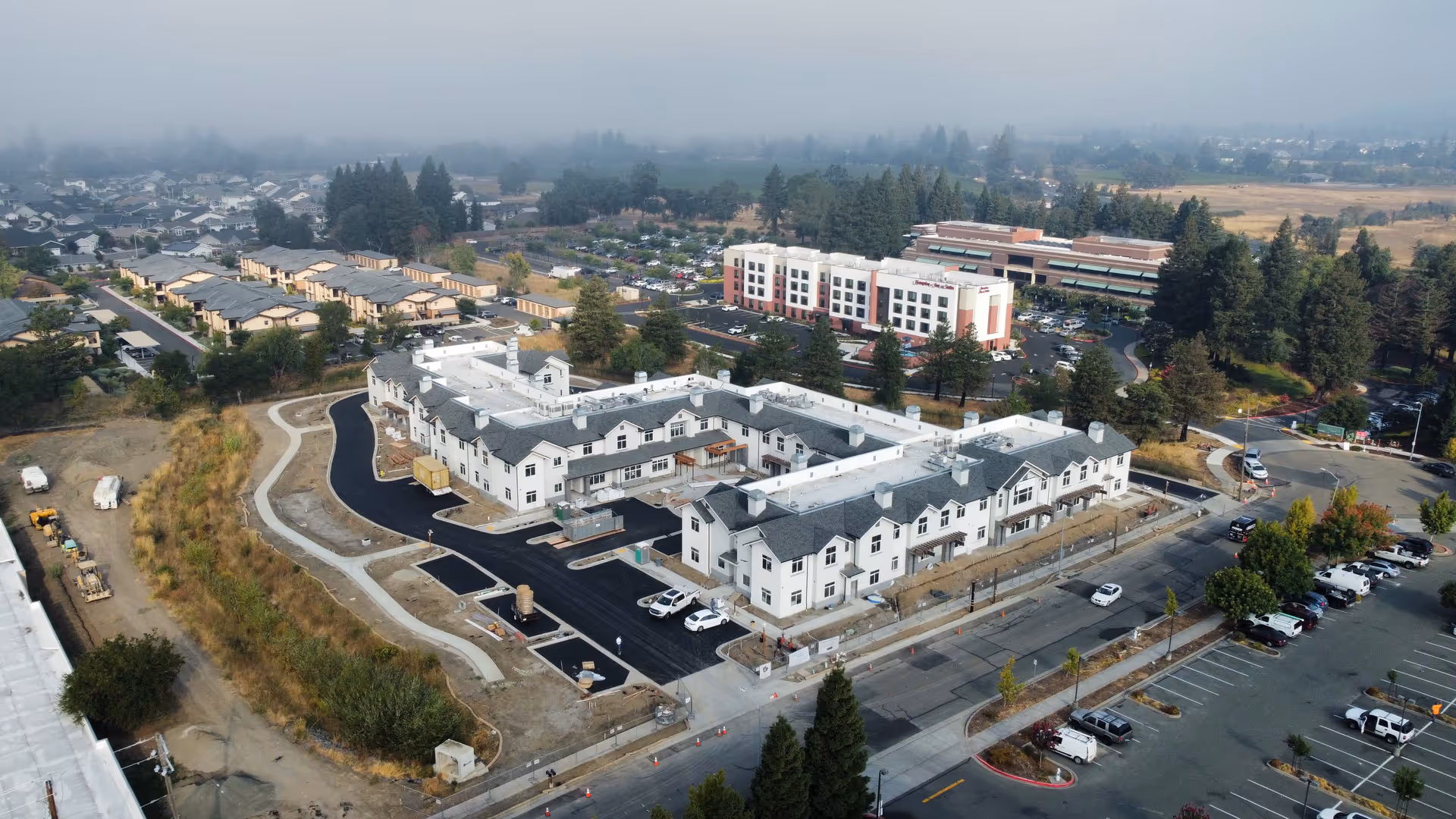 Aerial view of the Santa Rosa Hills Senior Living campus showing a U-shaped multi-story building under construction with parking lots, roads, and surrounding buildings and trees.