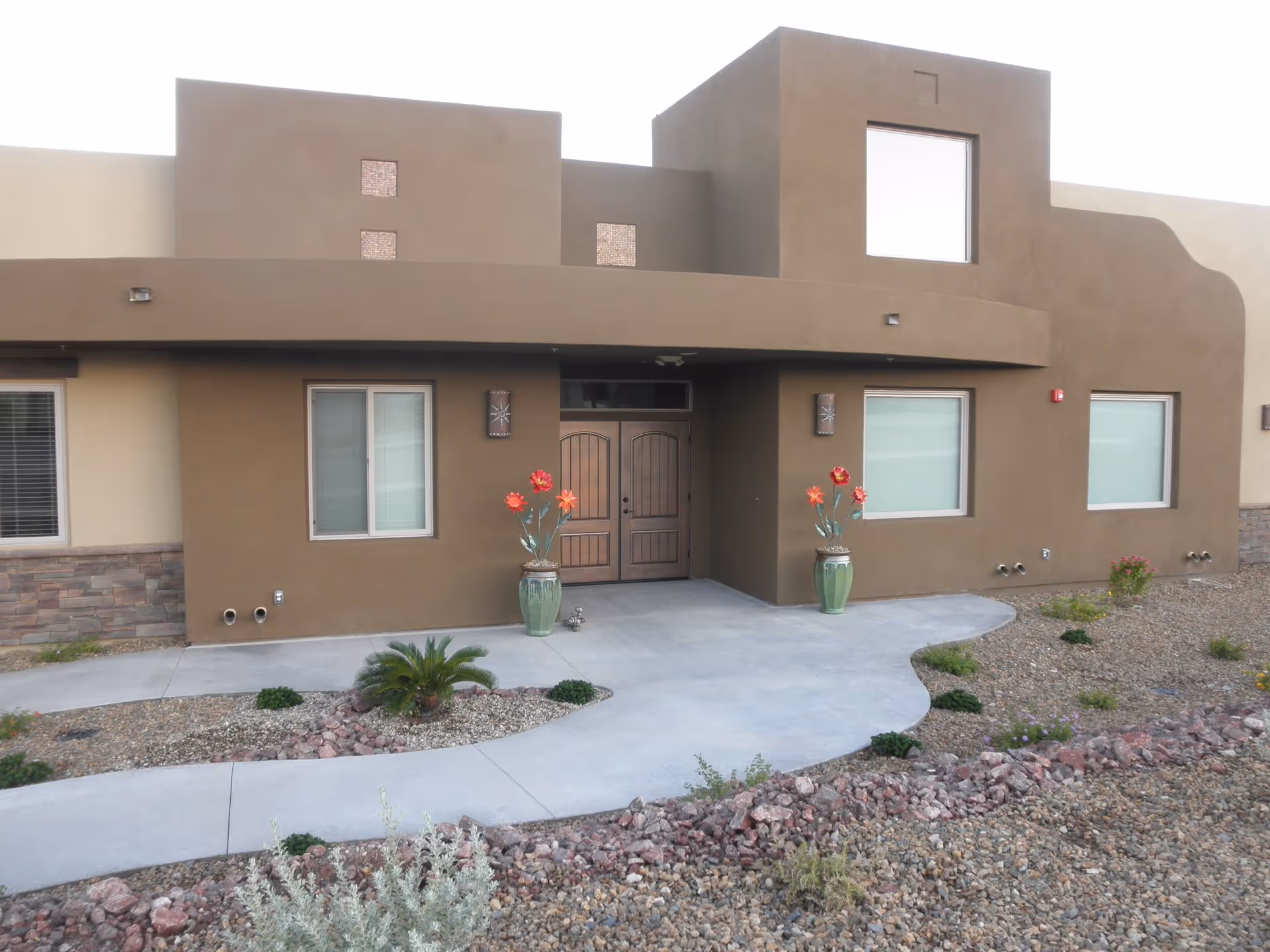 Exterior view of a modern building with brown stucco walls, two large wooden double doors at the entrance, and several windows with white blinds. The entrance is flanked by two large green pots with decorative red flowers. The surrounding landscape features a concrete walkway, gravel, small plants, and rocks.