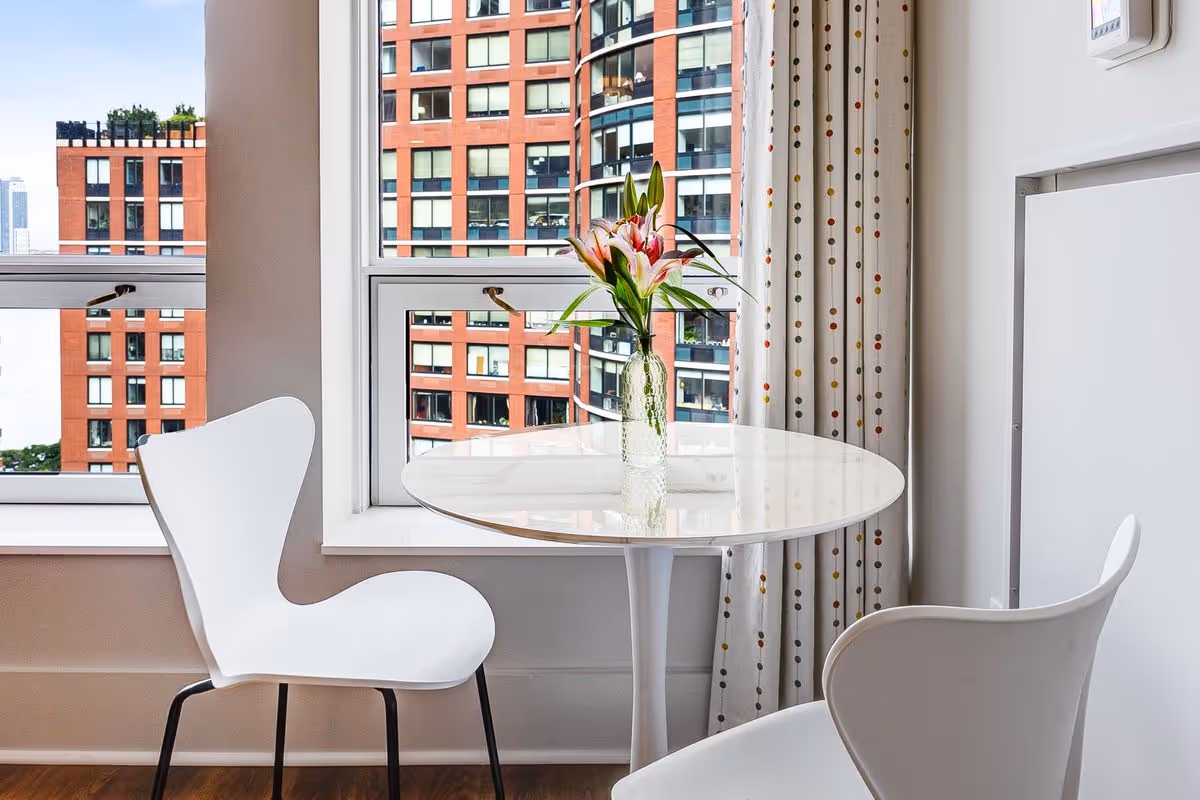 A small round white table with a glass vase holding pink lilies placed on top, positioned in front of a window with a view of red brick buildings outside. Two modern white chairs are placed around the table, and a curtain with a colorful dotted pattern hangs to the right side of the window.