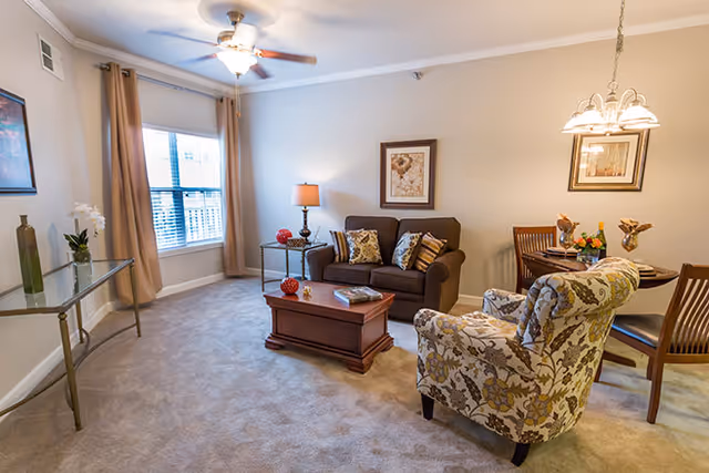 Comfortable living room with a sofa, patterned armchair, wooden coffee table, small dining table, and a window with curtains.