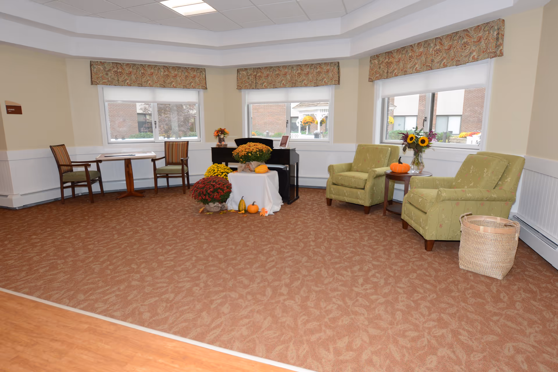 A cozy sitting area in a senior living facility with two green upholstered armchairs, a small round wooden table with a vase of sunflowers and a small pumpkin, a wicker basket, and a piano decorated with autumn flowers and pumpkins. The room has three windows with floral valances and beige walls with white wainscoting.