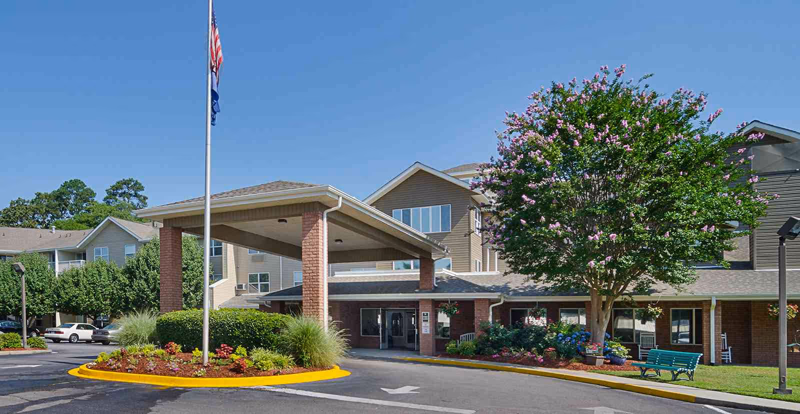 Exterior view of Holiday Columbia Pines senior living facility showing the main entrance with a covered drop-off area supported by brick columns, a flagpole with an American flag, landscaped flower beds, a large tree with pink flowers, and a green bench on the lawn under a clear blue sky.