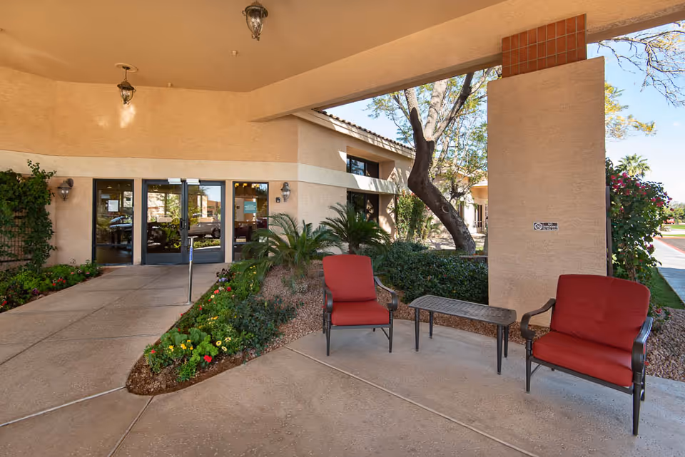 Covered outdoor entrance area of a building with two red cushioned chairs and a small table between them. There are plants and flowers along the walkway leading to glass double doors. The building has beige walls and a tiled roof, with trees and greenery visible in the background.