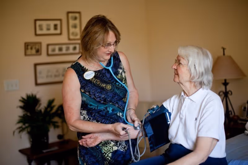 A healthcare professional uses a stethoscope and blood pressure cuff to check the blood pressure of an elderly woman seated on a bed in a warmly lit room with framed pictures on the wall and a lamp in the background.